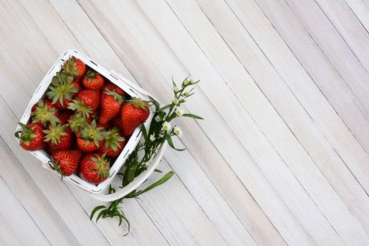 Overhead view of a basket of fresh picked strawberries on a white rustic wood table. A sprig of flowers is stuck between the handle and the basket. Horizontal format with copy space.