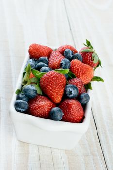 A bowl full of fresh blueberries and strawberries in a white rectangular bowl on a rustic wood kitchen table. Vertical format.