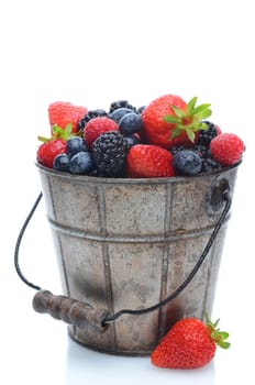 Assorted fresh picked berries in a pail with a wooden handle over a white background. Vertical format with reflection.