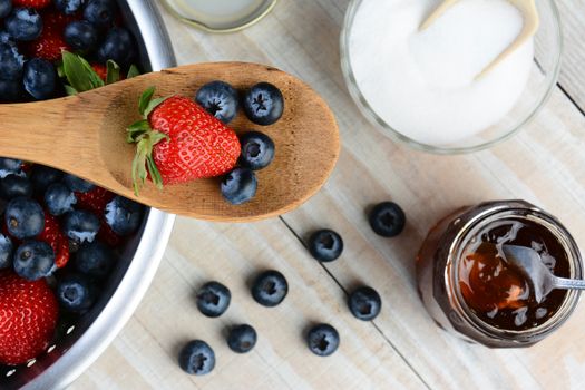 High angel photo of a Strawberry and Blueberries on a wooden spoon laying across a colander full of berries. A sugar bowl and jar of jam sitting on a rustic wooden farmhouse style table fill the background. Selective focus in horizontal format.