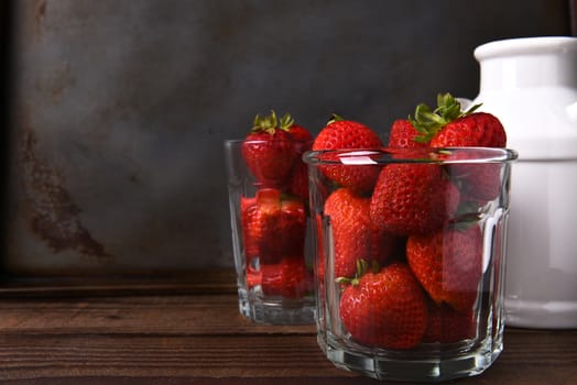 Horizontal still life with copy space of a glass filled with fresh picked strawberries on a wood table. In the background is a second glass of berries and a milk can.