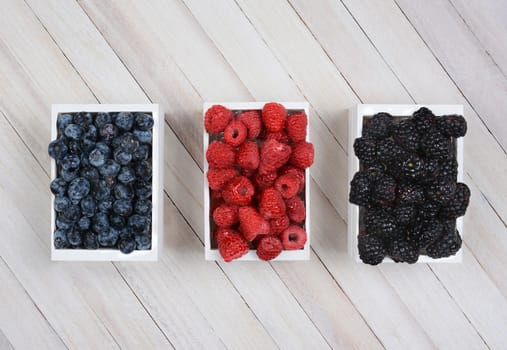 Three mini wood crates of berries on a rustic wood surface. Blueberries Raspberries and Blackberries in small white boxes shot from a high angle. 