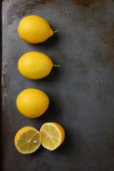 High angle shot of a group of lemons, three whole and one cut, on a metal baking sheet. Vertical format.