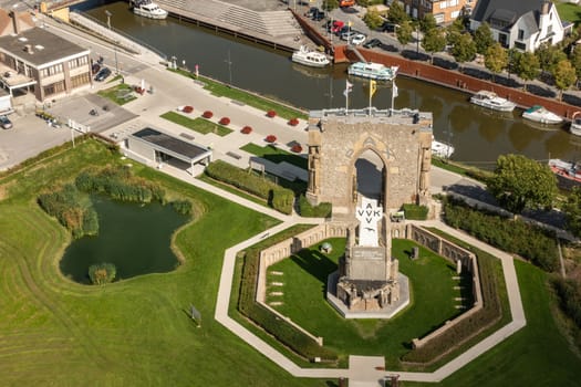 Diksmuide, Flanders, Belgium - September 15, 2018: Aerial view on PAX Gate and crypt ruin at IJzertoren plain with Ijzer River in back. AVV-VVK symbol on white cross, Green lawn, buildings and boats on dark water. Shot from top of IJzertoren.