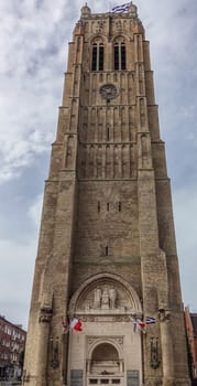 Dunkerque, France - September 16, 2018: the brown brick Belfry of Dunkirk towers over houses under cloudy blue sky. World wars memorial with flags at base.