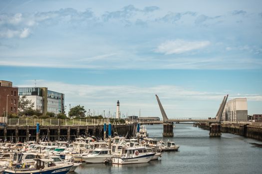 Dunkerque, France - September 16, 2018: The channel link to North Sea of old port in Dunkirk with motor yachts, bridge and buildings on side under light blue sky. Lighthouse on horizon.