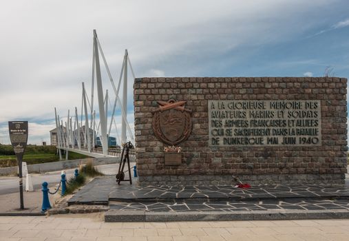 Dunkerque, France - September 16, 2018: Brown stone war memorial commemorating the Battle for Dunkirk in world war 2 near the beach. Pedestrial suspension bridge under blue-white sky.