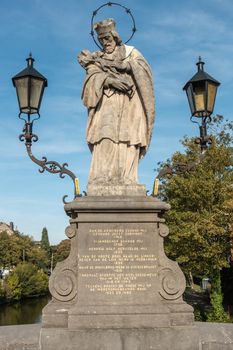 Kortrijk, Flanders, Belgium - September 17, 2018: Gray beige Statue of Saint John of Nepomuk on Bridge of Broel Towers under blue sky. Some green foliage in back and two lanterns.