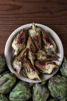 Grilled Artichokes: A plate of grilled artichoke pieces on a wood table with uncooked around the bottom of the plate. Vertical format.