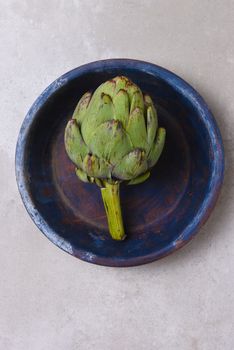 An Artichoke on a blue plate on light gray mottled background. Hig angle with copy space.
