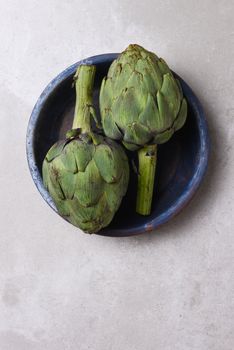 Two artichokes in a blue bowl on a mottled gray background with copy space.