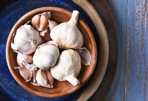 HIgh angle view of a bowl of garlic bulbs and cloves in a blow, on a rustic blue table. 