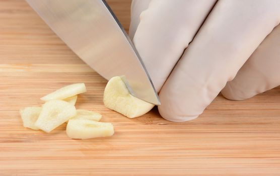 Closeup of a food service worker slicing a garlic clove on a wood cutting board. The chef is wearing a protective glove.