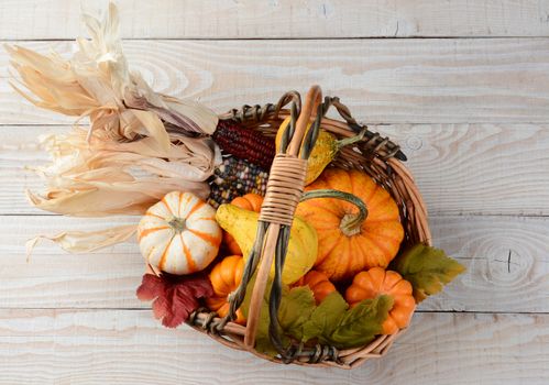 High angle view of an basket full of pumpkins, gourds, indian corn and leaves. The Autumn display is set on a rustic whitewashed wood table.