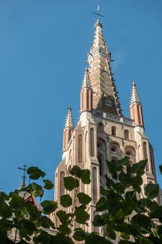 Brugge, Flanders, Belgium - September 19, 2018: Spire of brick Notre Dame Cathedral tower against blue sky with dark green foliage up front in Bruges.