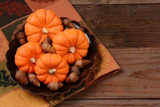 Overhead view of a bowl full of mini pumpkins and acorns on a fall tablecloth and rustic wooden table. Horizontal format with copy space.