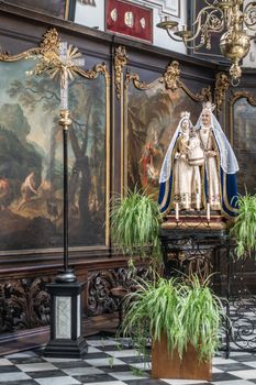 Brugge, Flanders, Belgium - September 19, 2018: Wall covered in paintings with gold decorations behind statue of Saint Anna and the Madonna in Saint Anna Church in Bruges. Some green plants up front.