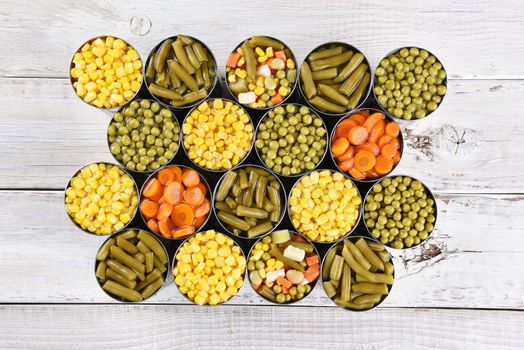 High angle shot of a group of canned vegetables on a rustic white wood table. Several varieties of opened cans including, corn, green beans, peas, carrots and mixed veggies.