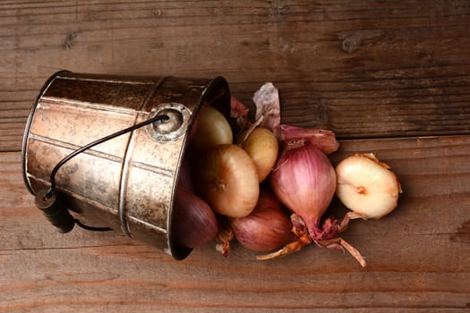 Overhead view of onions and shallots spilling from a bucket onto a rustic wooden table.