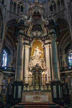 Ghent, Belgium - September 23, 2018: Main altar with statue set in chancel of Saint Bavo Cathedral. Light falss on white statue framed in gold. Rest mainly in gray stone and darker environment.