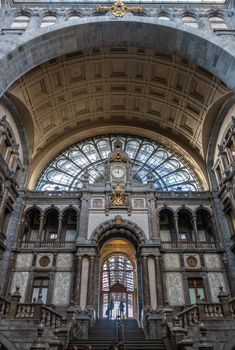 Antwerp, Belgium - September 24, 2018: Gray, beige, brown Monumental hall and arch over stairway to train hall at Antwerpen Centraal railway station with golden decorations and clock.