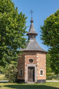 Bokrijk, Belgium - June 27, 2019: Red brick octagonal chapel of Metsteren with dark gray roof and cross on top surrounded by trees. Brown door is closed.