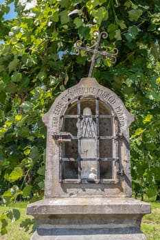 Bokrijk, Belgium - June 27, 2019: Closeup of small gray stone chapel niche along road with white statue of Virgin Mary. Iron cross on top is slanted. Green foliage in back.