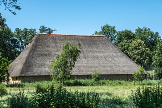 Bokrijk, Belgium - June 27, 2019: Giant barn with gray straw roof is set in green of meadow and surrounding trees under blue sky.