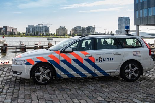 Amsterdam, the Netherlands - June 30, 2019: Closeup of whtie-blue-red police station wagon at IJDock unde rmorning light. Outlet to sea and high rise buildings in back under light blue sky.