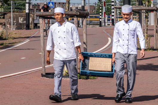 Amsterdam, the Netherlands - June 30, 2019: Two young male kitchen helpers in uniform carry blue crate with Lamb meat box along street near IJdok.