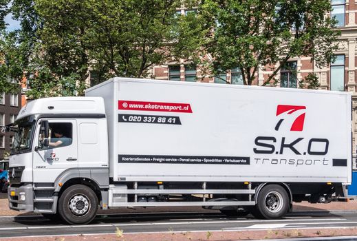 Amsterdam, the Netherlands - June 30, 2019: Large white transport truck with black and red lettering for S.K.O. company. Green foliage above. Brown-white facades in back.