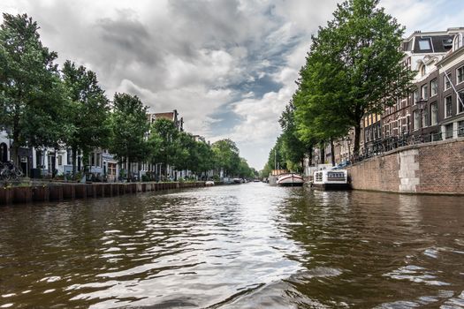 Amsterdam, the Netherlands - June 30, 2019: gray white cloudscape with blue patches above Prinsengracht canal with green trees along the walls. Some boats and houses.