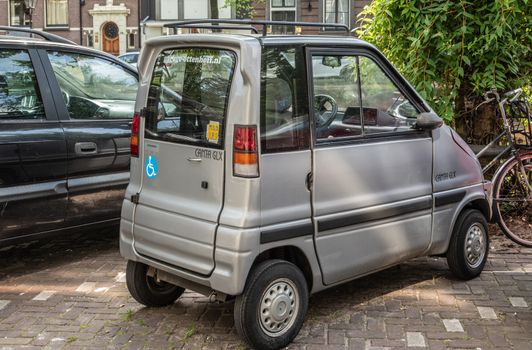 Amsterdam, the Netherlands - June 30, 2019: Small gray painted mini car called Ganta GLX  parked along canal . Green foliage.