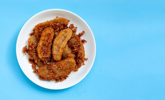 Fried bananas with sesame seeds on white ceramic round plate on blue background. Top view with copy space