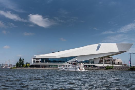 Amsterdam, the Netherlands - June 30, 2019: White Eye Film Museum under blue sky with some white clouds. IJ water in front. Pleasure boat passing by.