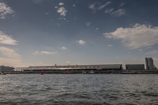 Amsterdam, the Netherlands - June 30, 2019: Terminal hall of Centraal Railway Station seen from IJ water under blue sky with white clouds. Other buildings at the edges.