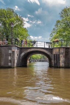 Amsterdam, the Netherlands - June 30, 2019: Small canal bridges under gray-blue-white cloudscape with green foliage and people on land. Seen from canal water.