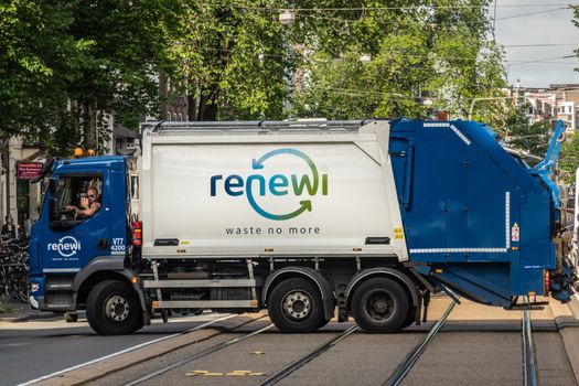 Amsterdam, the Netherlands - July 1, 2019: Blue and white garbage for recycling collection truck under green foliage in street scene. Driver active. Name on truck is Renewi.