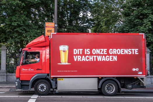 Amsterdam, the Netherlands - July 1, 2019: Red Amstel beer delivery truck although it claims to be the greenest truck. Driver active. Image of full Amstel bier glass. Green foliage.