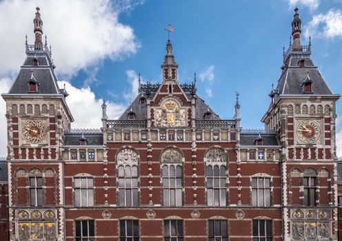 Amsterdam, the Netherlands - July 1, 2019: Red brick with white trim upper part facade Centraal Railway Station with towers, clocks, and plenty of frescoes under blue sky with white clouds.