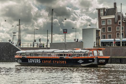 Amsterdam, the Netherlands - July 1, 2019: Lovers Canal Cruises boat on the move in canal under heavy cloudscape on top of dark water. Street with houses and people in back.