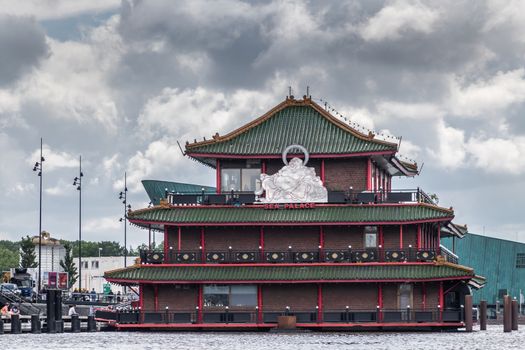 Amsterdam, the Netherlands - July 1, 2019: Red-green-brown short facade with huge white Buddha statue of Sea Palace floating restaurant at Oosterdokskade under heavy cloudscape.