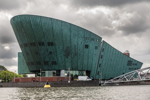 Amsterdam, the Netherlands - July 1, 2019: Modern boat hull building housing the NEMO science museum on border of Oosterdok in black water under havey rainy cloudscape.