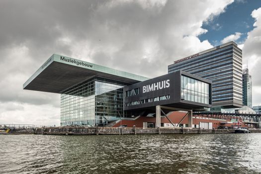 Amsterdam, the Netherlands - July 1, 2019: Modern cubic building housing the music theater and school with Jazz Bimhuis on platform in black water IJ river under havey rainy cloudscape.