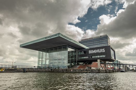 Amsterdam, the Netherlands - July 1, 2019: Modern cubic building housing the music theater and school with Jazz Bimhuis on platform in black water IJ river under havey rainy cloudscape.