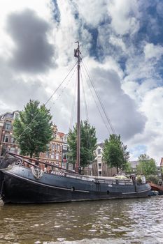 Amsterdam, the Netherlands - July 1, 2019: Avance sailing houseboat to live in on canal under heavy cloudscape. Some green foliage.