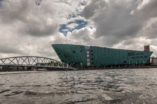 Amsterdam, the Netherlands - July 1, 2019: Modern boat hull building housing the NEMO science museum on border of Oosterdok in black water under havey rainy cloudscape.
