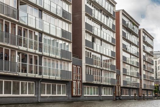 Amsterdam, the Netherlands - July 1, 2019: Line of simple, modern apartment buildings with lots of glass windows dropping down into canal under cloudscape.