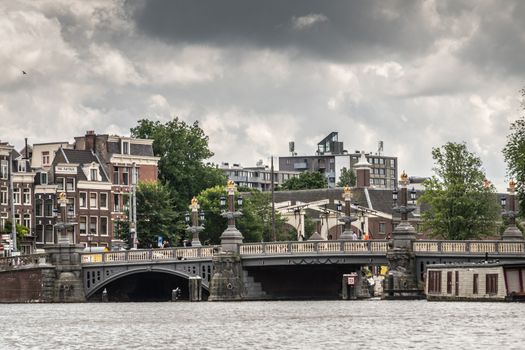 Amsterdam, the Netherlands - July 1, 2019: Blauwbrug, bridge over Amstel River under heavy cloudscape. Rows of houses, green foliage and white frame of Walter Suskind bridge.