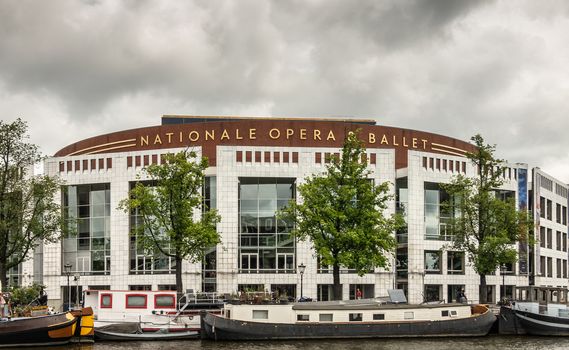 Amsterdam, the Netherlands - July 1, 2019: Large white building with brown crown of National Opera and Ballet along Amstel River with houseboats in front. Golden letters on top. Cloudscape.